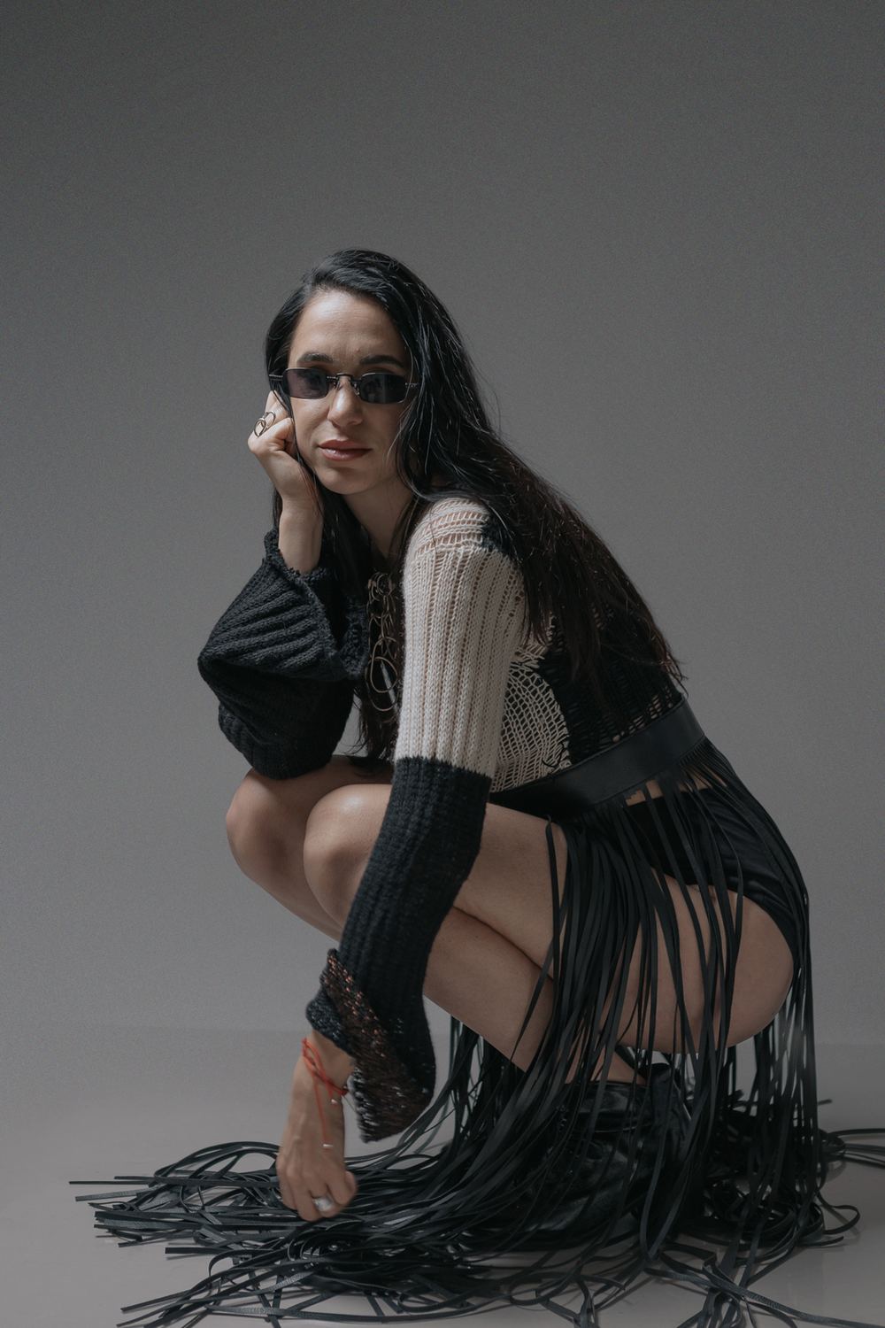 Crouching in a knit top and long fringe skirt, grey backdrop